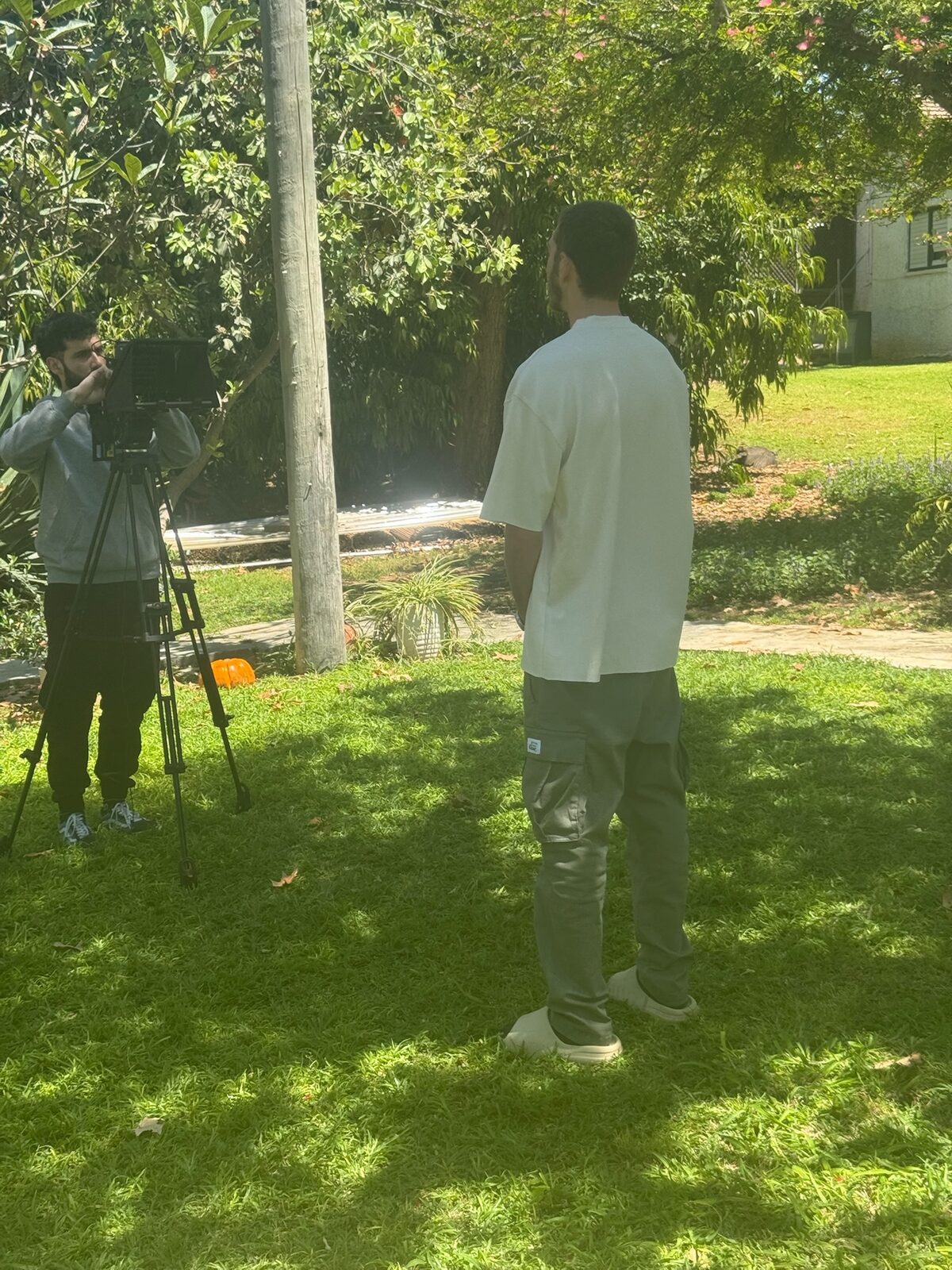 מאחורי הקלעים של יום צילום בחוץ, צלם וידאו עם מצלמה על חצובה מתעד מרואיין על רקע מדשאה ירוקה וצמחייה.