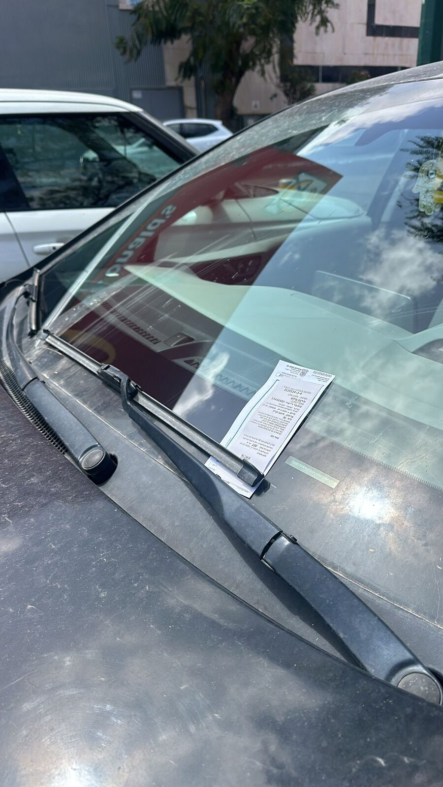 דוח חניה עירוני מונח מתחת לווישר על שמשת רכב מאובק, ברקע נראית סביבה אורבנית.